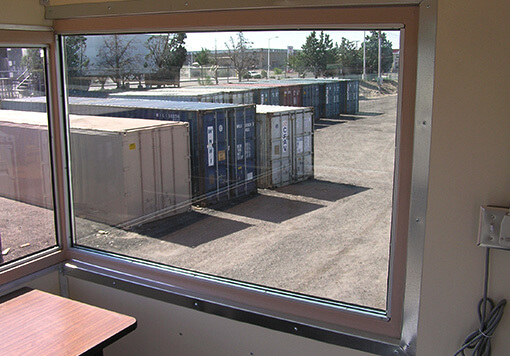 a window inside a storage container, outside the storage container are nine more containers in different colors, there are also some trees outside.