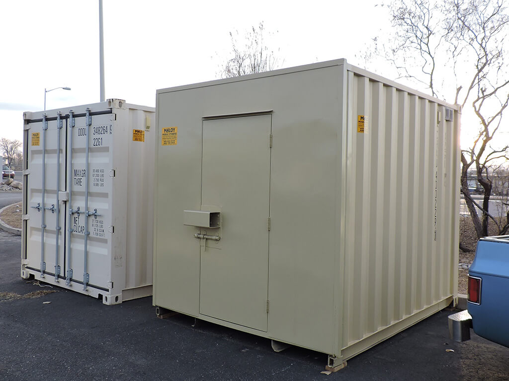 two 10ft storage container, a gold and white storage containers, both have a small yellow stickers saying Maloy Mobile Storage and the company contact number, behind the containers are some dead trees.